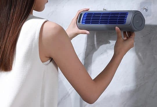 Grey FreshAir Pro long tube fan with a blue slatted grille being hung on a wall hook by a woman in a white top