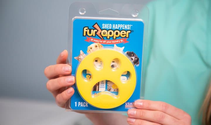 Hand holding a FurZapper retail blister pack with a yellow disc and Shed Happens branding