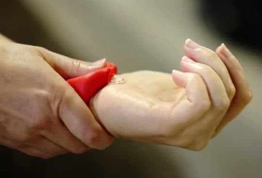 Close-up of fingers pressing a red Handsan Wrist silicone band so a drop of clear sanitizer comes out onto the other palm