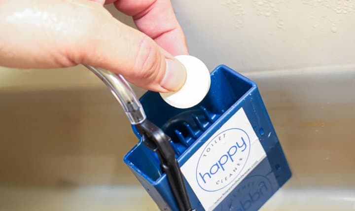 Hand dropping a white cleaning tablet into the Happy Toilet Cleaner holder inside a toilet tank