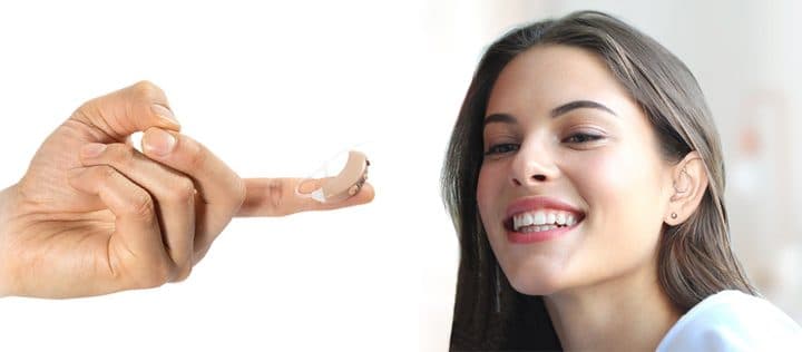 A small beige behind-the-ear HearingAid Max amplifier balanced on a fingertip, next to a smiling woman with long dark hair