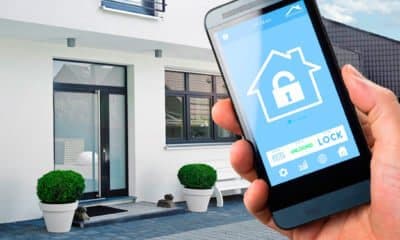 Hand holding a phone showing a house lock icon in front of a modern house, illustrating the HomeNETIX Home and Away Mode add-on
