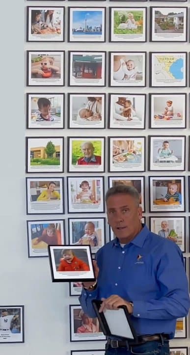 Man in a LifeVac shirt standing beside a wall of framed photos of people the brand says were saved