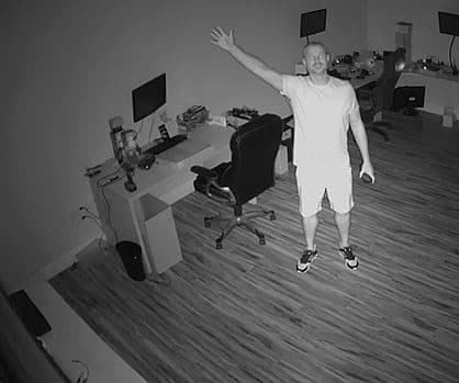 Black and white infrared view of a man waving in a home office with desks and monitors