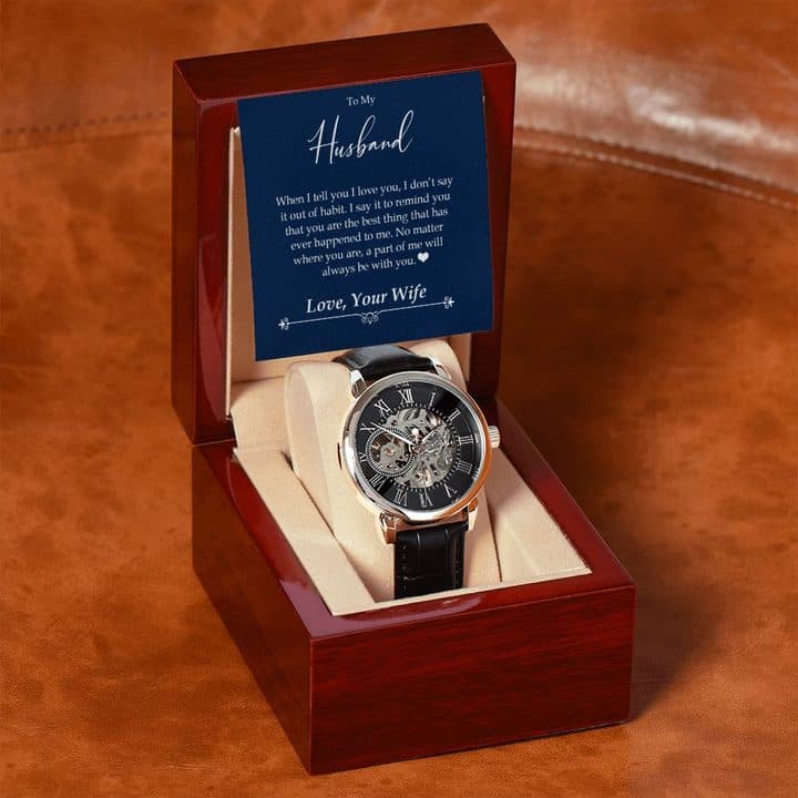 Rose gold-tone openwork watch on a cream cushion in a mahogany-style box on a tan leather sofa, with a To My Husband card signed Love, Your Wife