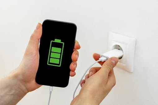 Hand plugging a white USB power adapter into a wall socket while charging a phone with a green battery icon on screen