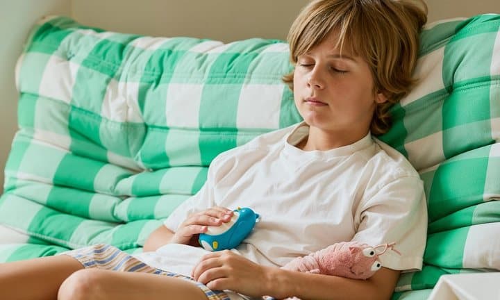 Boy sitting on a sofa holding a blue Moonbuddy bird in his lap with his eyes half closed