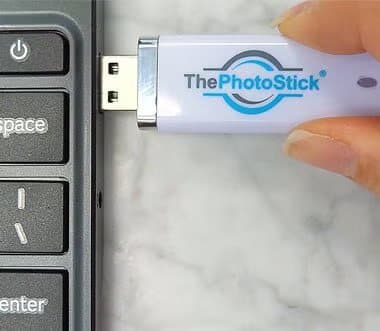 Hand plugging a white ThePhotoStick into the USB port of a laptop