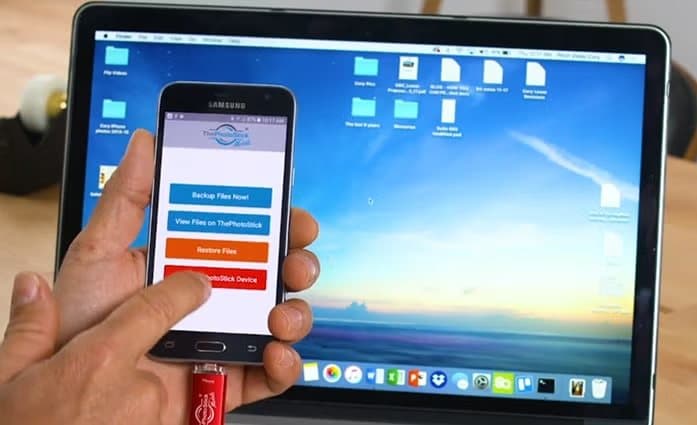 Phone running ThePhotoStick Mobile app with backup buttons, held in front of a laptop screen
