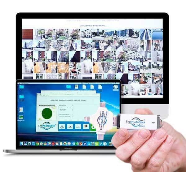 Hand holding ThePhotoStick Omni beside a laptop running its backup program, with photo thumbnails on a monitor behind