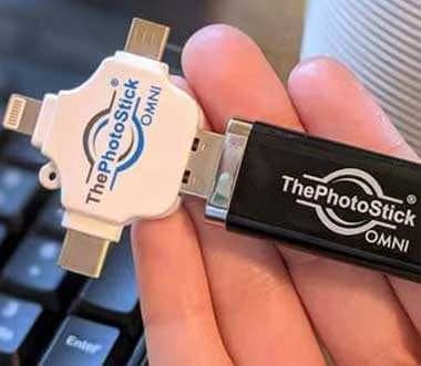 Hand holding ThePhotoStick Omni with its white adapter showing Lightning, USB-C and micro-USB plugs