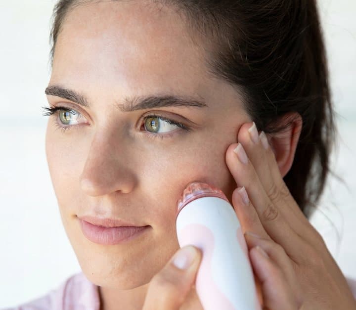 Woman gliding a white and pink PMD microderm device across her cheek