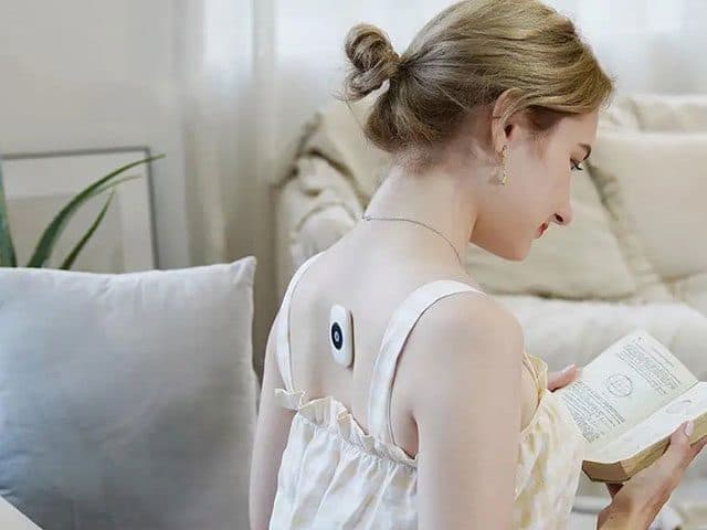 Woman reading a book on a sofa with the small white PostureRemind Pro attached to her bare upper back