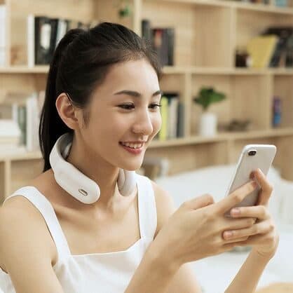 Smiling woman wearing a white SmartRelief Pro around her neck while looking at her phone, sitting by bookshelves