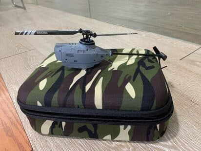 Grey mini helicopter resting on top of a green and brown camouflage hard-shell case on a wooden floor