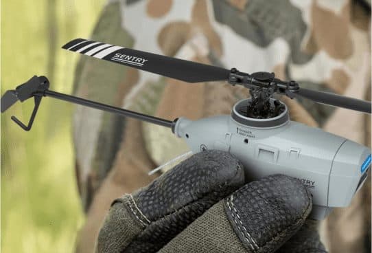Gloved hand holding a small grey helicopter with a long tail boom and two black rotor blades printed Sentry