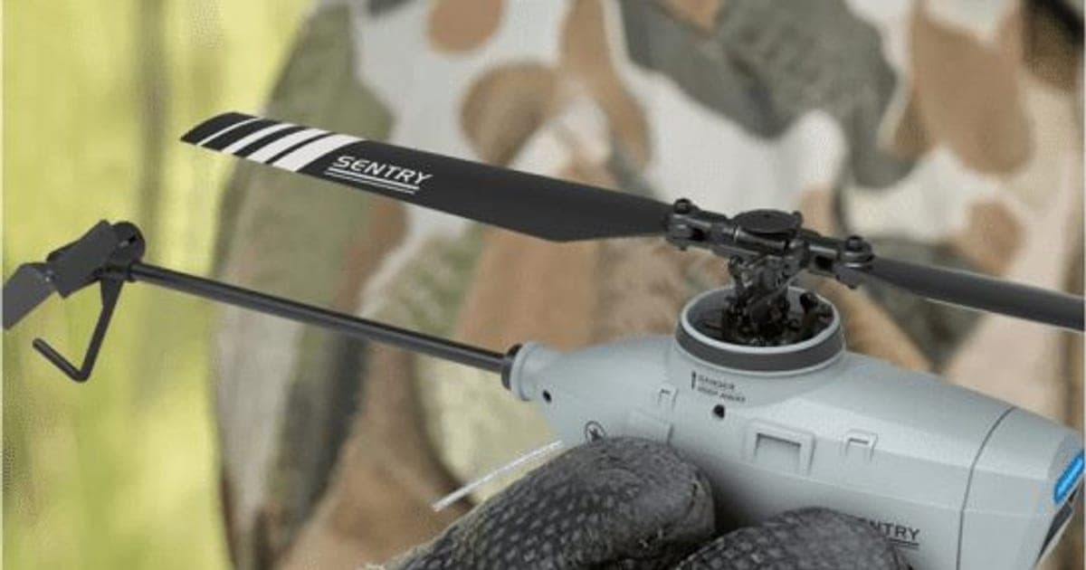 Grey StealthHawk Pro mini helicopter with long black rotor blades marked Sentry, held in a gloved hand against camouflage fabric
