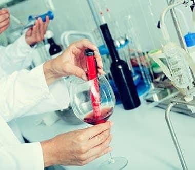Person in a lab coat pouring red wine into a glass in a laboratory