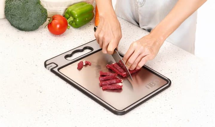 Hands slicing raw beef on the metal side of a TIBO cutting board beside peppers and broccoli