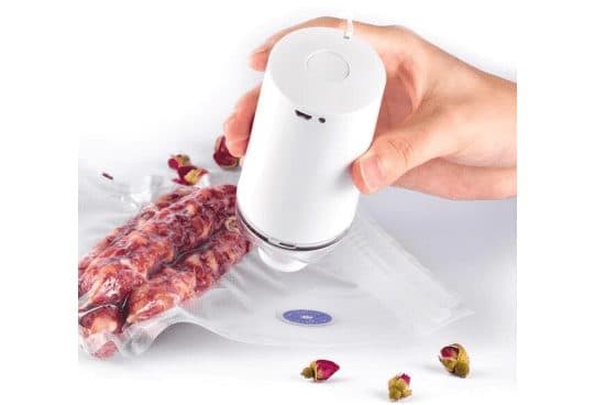 White cylindrical Vacuumer Max pump pressed onto the blue valve of a vacuum bag holding sausages, with dried rosebuds scattered on the table