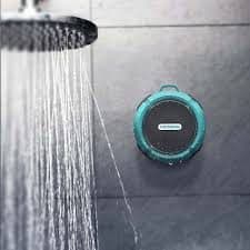 A teal round shower speaker stuck to a grey tiled wall next to a running shower head, used for the WaterBOOM 360 suction cup add-on