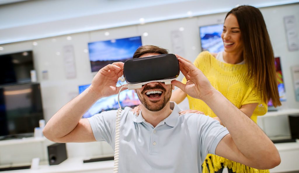 Young satisfied happy handsome man testing VR goggles while his smiling girlfriend standing behind and supporting him in a tech store.